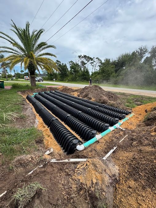 Black corrugated drainage pipes laid in a trench, with green and white pipes, dirt, and a palm tree.