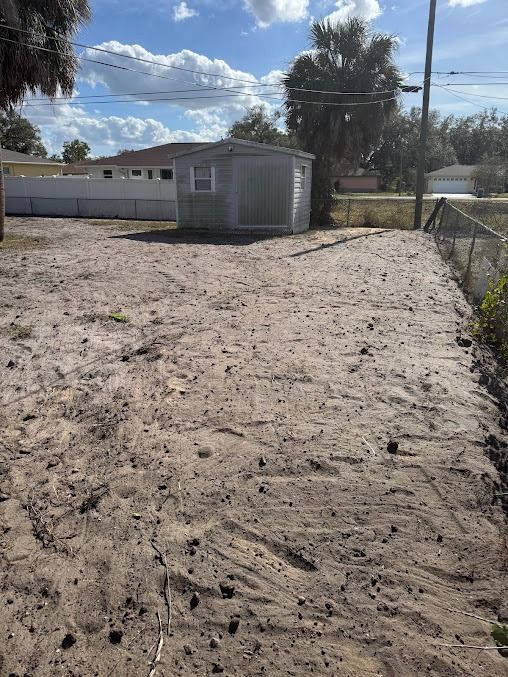 A sandy, empty backyard with a small shed and a chain-link fence. Blue sky and clouds overhead.