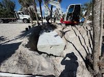 Excavator and truck near a partially buried concrete tank in sandy soil, with palm trees and a sunny sky.