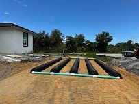 A septic system installation with black effluent tubes and a green pipe, in a sandy field, near a house.