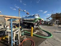 A tanker truck with a large hose connected, likely pumping liquid, on a concrete surface near a facility.