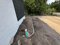 A green PVC pipe emerges from a trench in dirt next to a building with black siding and white stucco.