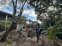 Construction site with workers, excavator, and large hole near a building and trees on a cloudy day.