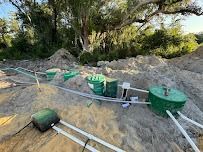 Green septic tanks and pipes on sandy ground, with trees in the background.