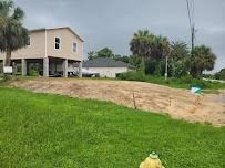 House on stilts beside a grassy lot with utility poles and trees under a cloudy sky.