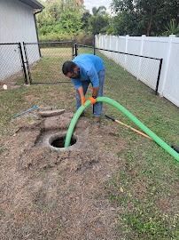 Man in blue shirt and jeans, pumping a green hose into an open septic tank in a yard.