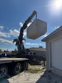 A crane lifting a large, light-colored object near a garage on a sunny day.