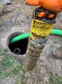 A person in orange gloves holds a clogged septic tank filter over an open tank. Green hose visible.