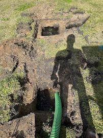 A person's shadow cast over a hole dug in the ground with a green hose nearby.