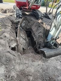 Excavator digging a trench in the dirt; red and white vehicle, grey soil.