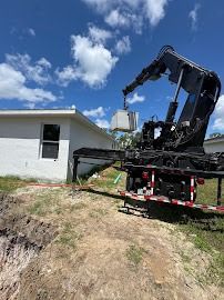 A crane lifting a large, square object near a white house on a sunny day.