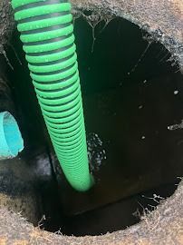 A close-up view inside a dark septic tank. A green corrugated hose and a blue pipe are visible.