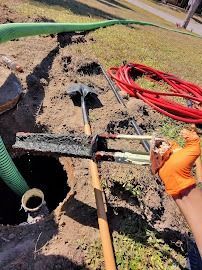 Construction site with exposed pipes, orange-gloved hand, shovels, and red and green hoses.