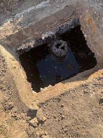 Open square pit in dirt; dark water surrounds a pipe.