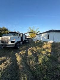 White flatbed truck parked on overgrown grass near a white building under a blue sky.