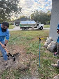 Man digs with shovel; a water truck and bags of material are in the background, outdoors.