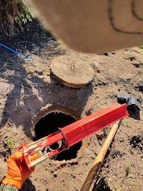 Person holding a red tool over an open manhole. Dirt surrounds the opening, and a lid rests nearby.