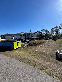 A construction site with a truck and dumpster. Green grass, blue sky, and houses in the background.