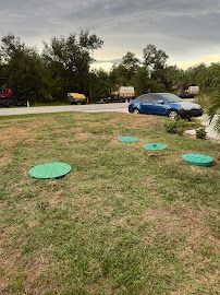 Lawn with green septic lids. A blue car is parked on a driveway. Several vehicles are parked on a road lined with trees.
