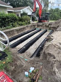Excavated yard with black drainage pipes installed for a septic system; excavator in background.