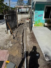 Trench dug for pipes along a building; white pipe laid in trench, person's shadow visible.