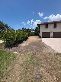 A driveway next to a building with two garages, with a brown area of soil in the foreground, and shrubs to the left.