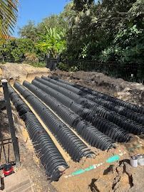 Black corrugated pipes laid in trenches, likely for drainage, in an outdoor setting with trees in the background.