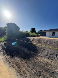 Sunlit construction site, dirt ground, yellow caution tape, green and white box, partially built house, bright blue sky.