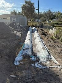 Trenches with white wrapped pipes in a residential yard, under construction, with a shed in the background.