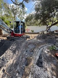 Mini excavator digging a trench in a yard near a white fence, under a blue sky.
