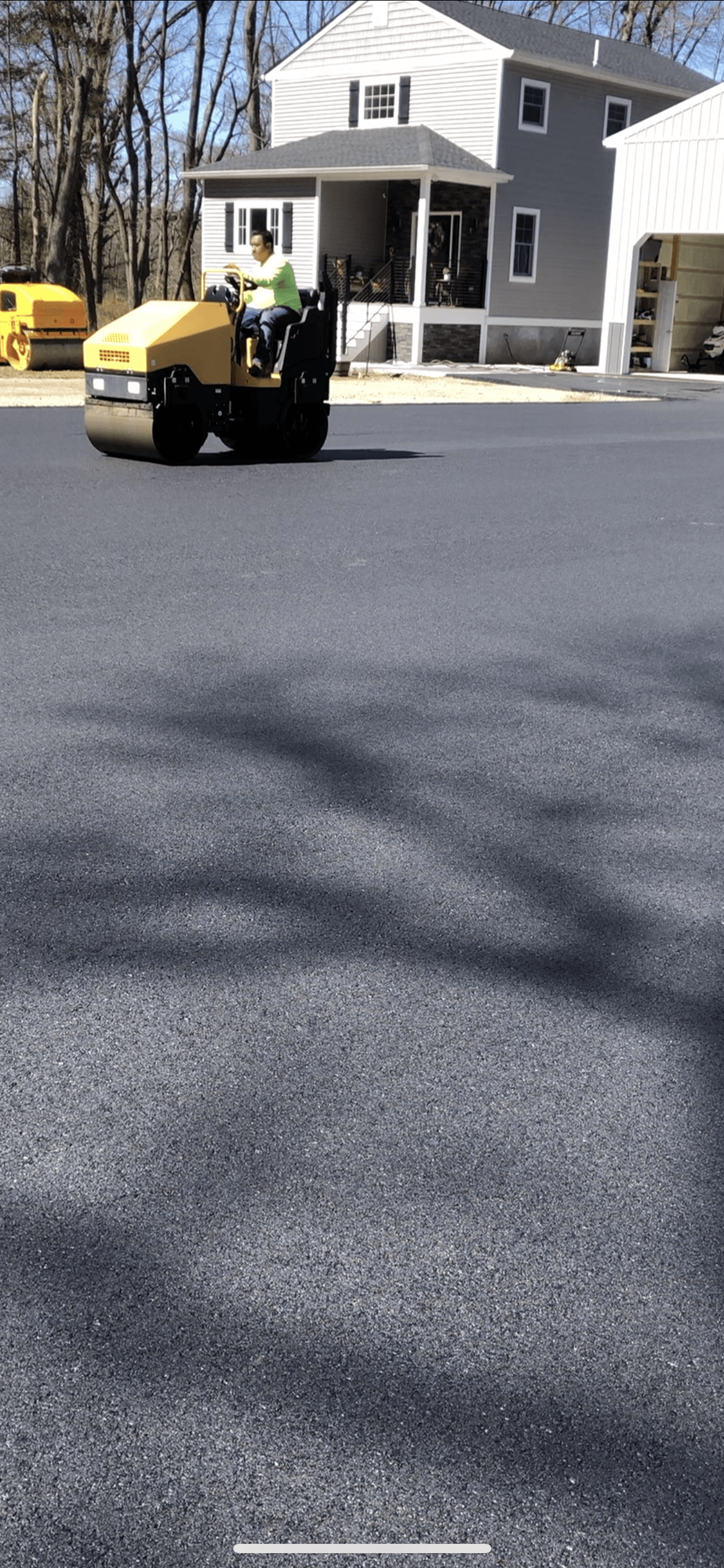 A yellow roller is rolling asphalt on a road in front of a house