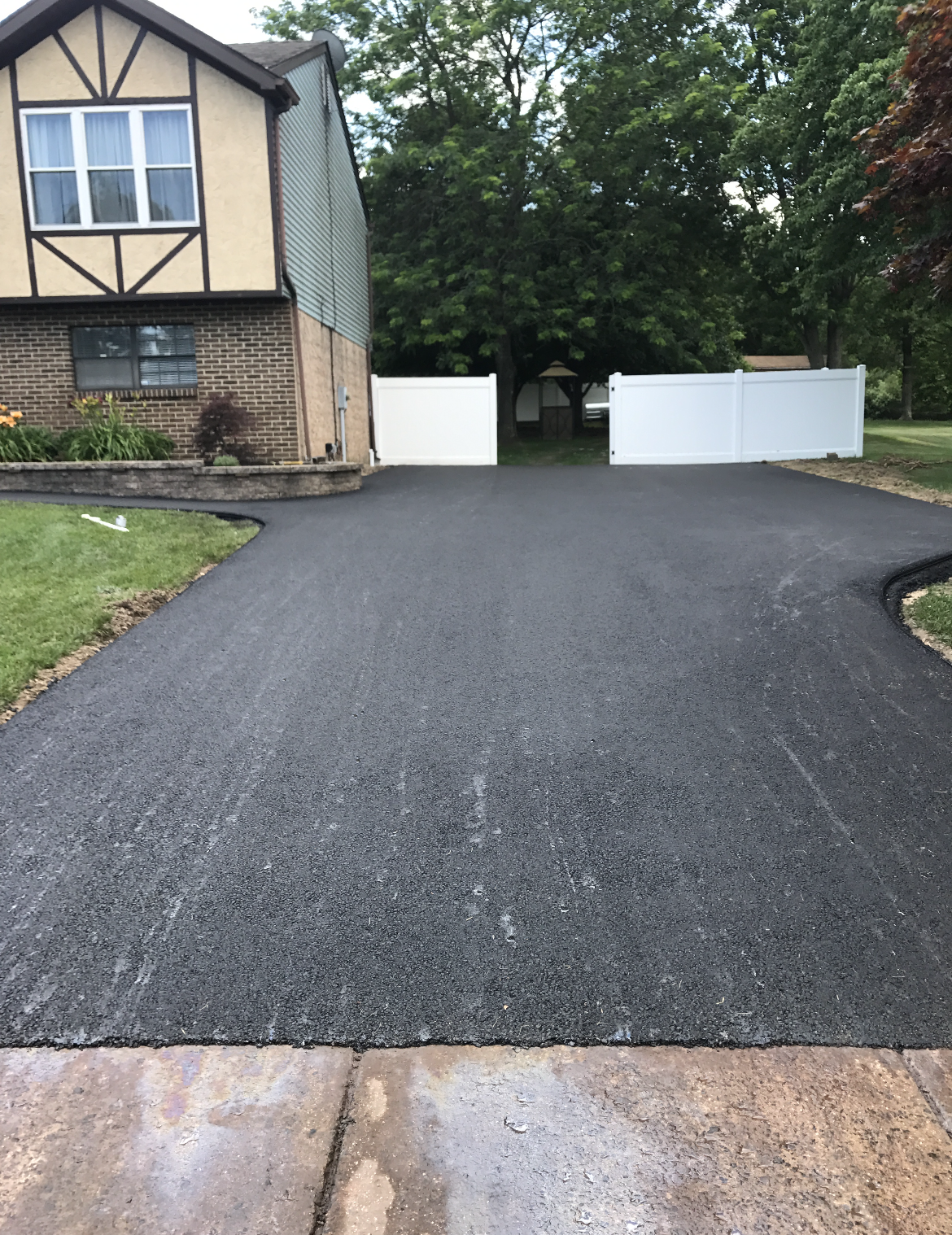 An asphalt driveway leading to a house with a white fence