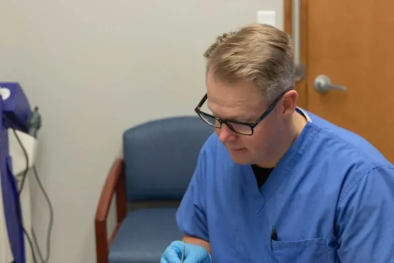 Man in blue scrubs and glasses looking down, possibly examining something in a medical setting.
