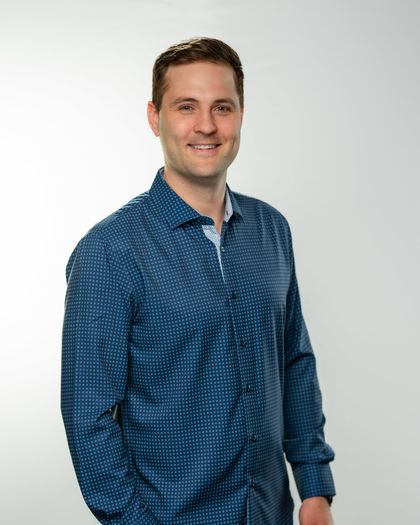 A smiling man in a white shirt and blue tie, against a gray background.