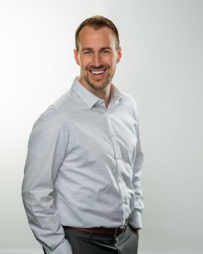 Smiling man in a gray suit jacket, white dotted shirt, and dark hair against a white background.