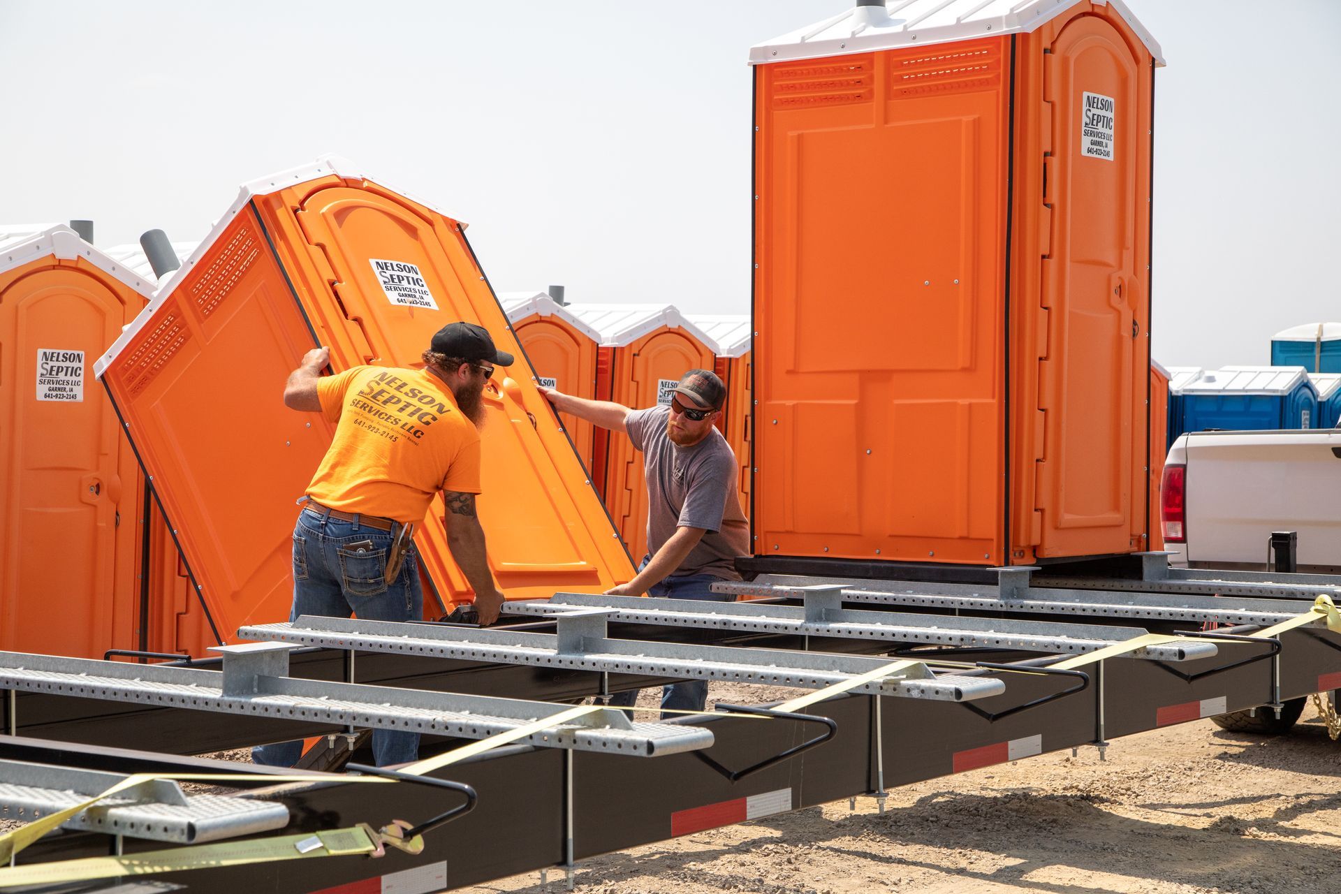 Two men are working on a trailer with portable toilets in the background