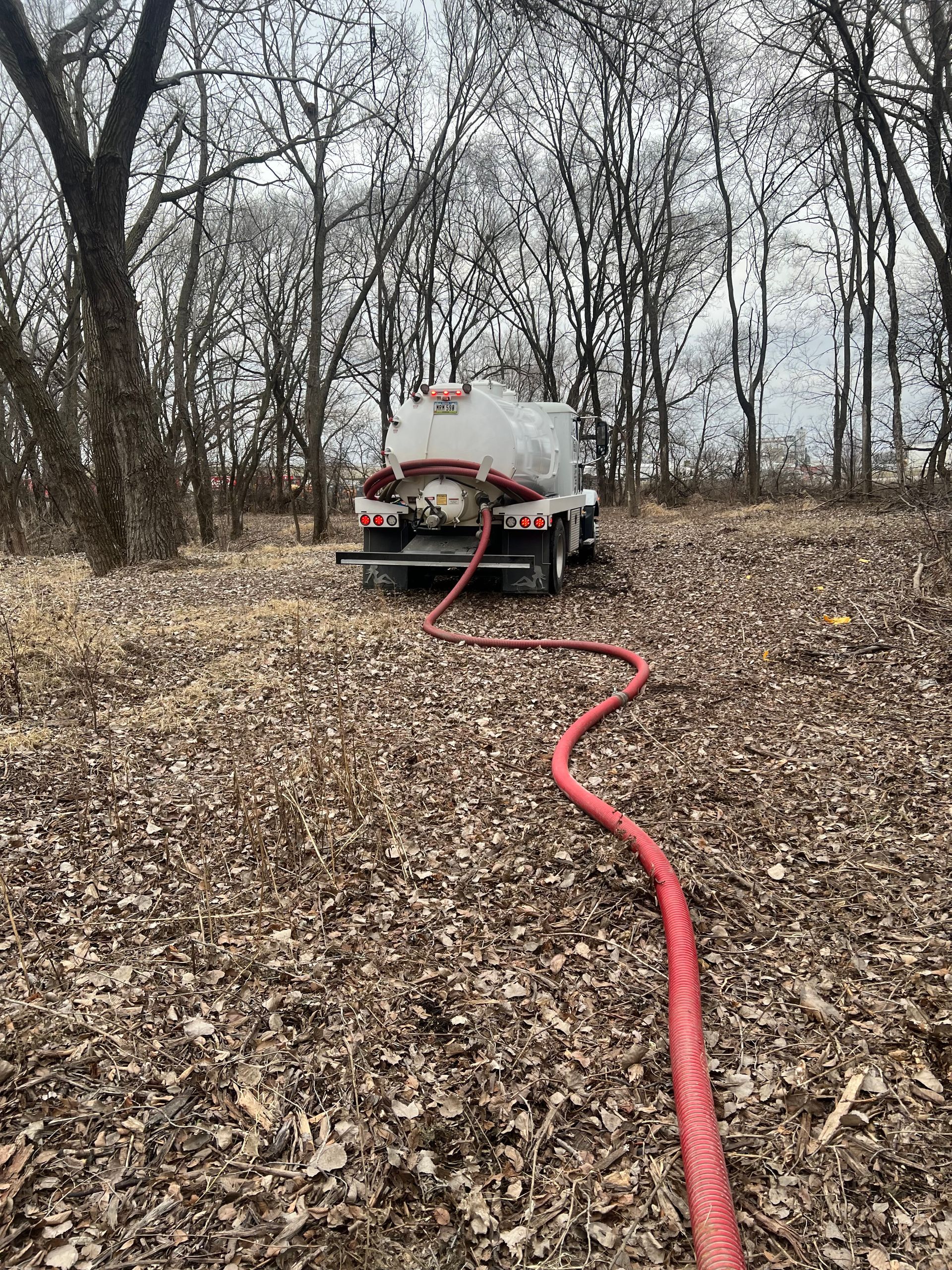 A vacuum truck with a red hose attached to it is sitting in a field of leaves