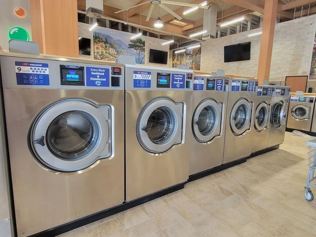 Row of stainless steel washing machines in a bright laundromat, with blue digital display panels.