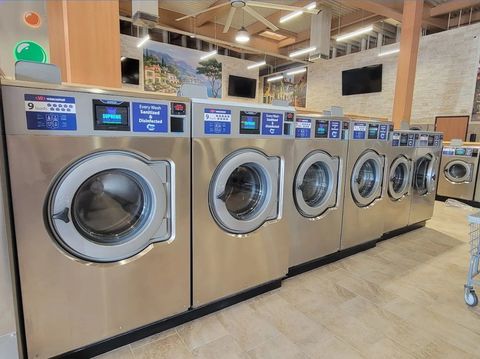 Row of stainless steel front-load washing machines in a laundromat with blue digital displays.