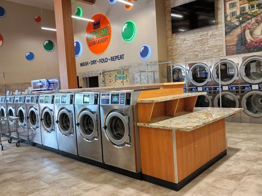 Row of silver washing machines in a laundromat with orange counters and colorful wall decor