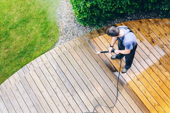 Person pressure-washing a wooden deck beside green grass and a gravel path