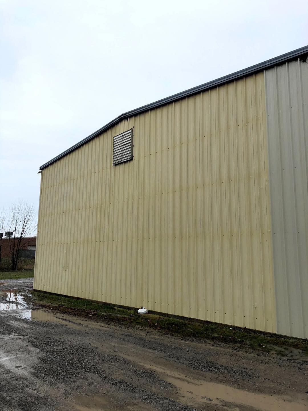 Side of a beige metal building with a small window beside a muddy gravel road under an overcast sky