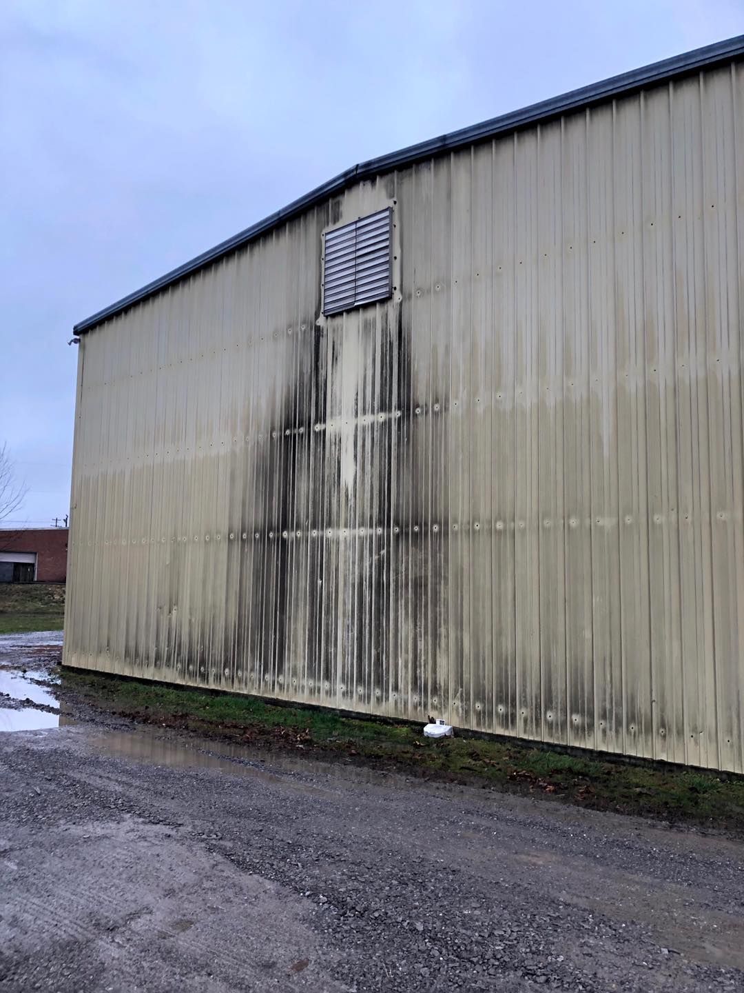 Stained beige warehouse wall with a vent, beside a wet gravel lot under a cloudy sky