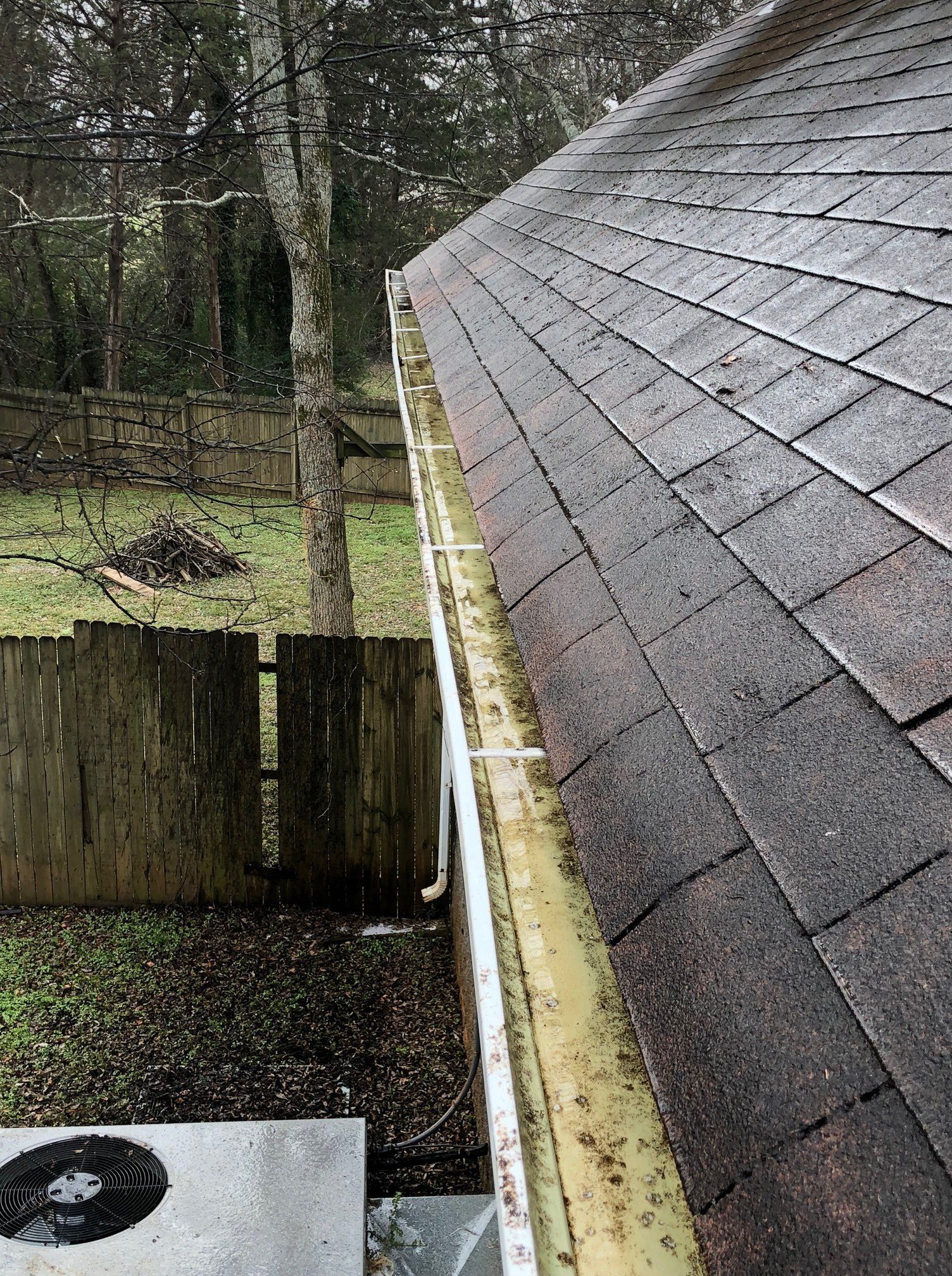 View from a roof edge with wet asphalt shingles, a gutter, and a fenced yard below amid trees