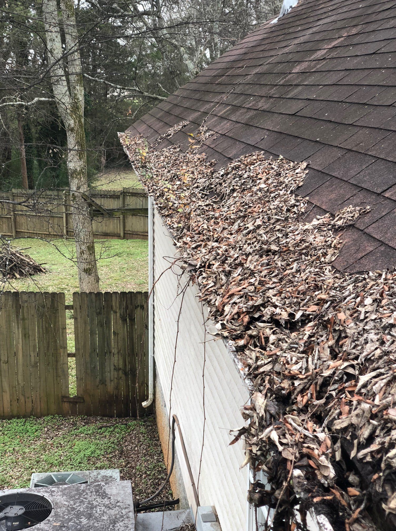 Gutter packed with leaves along a house roof, beside a fenced backyard and trees