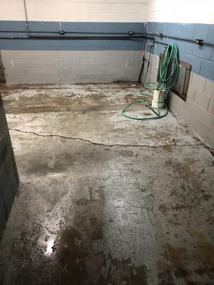 Empty concrete basement with wet floor, cinderblock walls, and a green hose on the right.