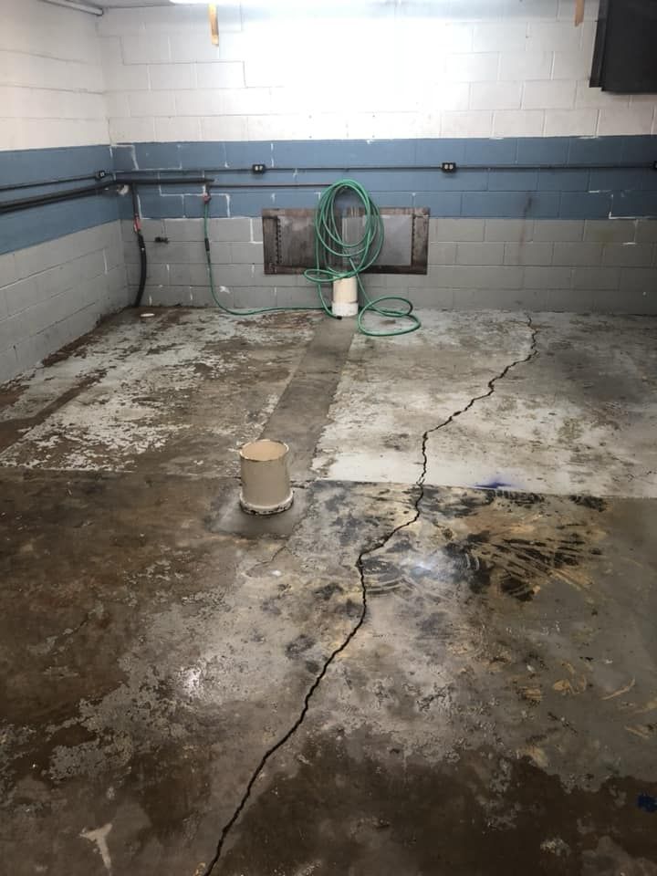 Dilapidated basement room with cracked concrete floor, damp stains, and a green hose on the back wall.