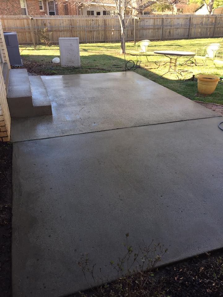 Concrete backyard patio with steps, grass, wooden fence, and a yellow bucket in the sun
