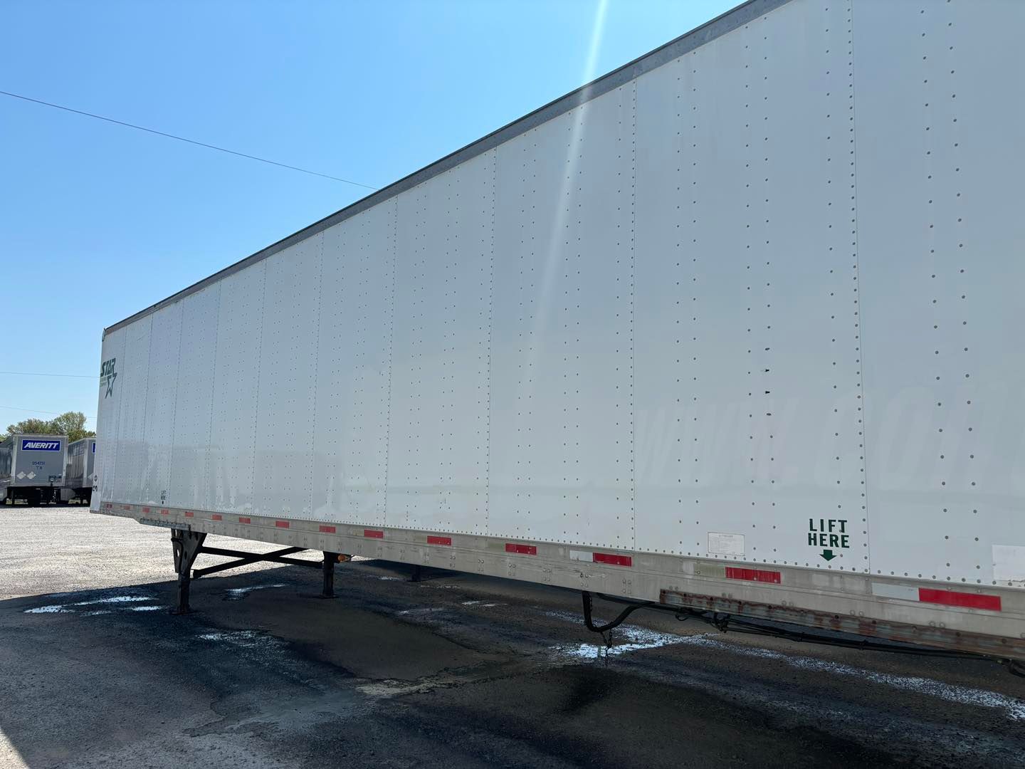 White semi-trailer parked on a gravel lot under a blue sky, with another truck in the distance