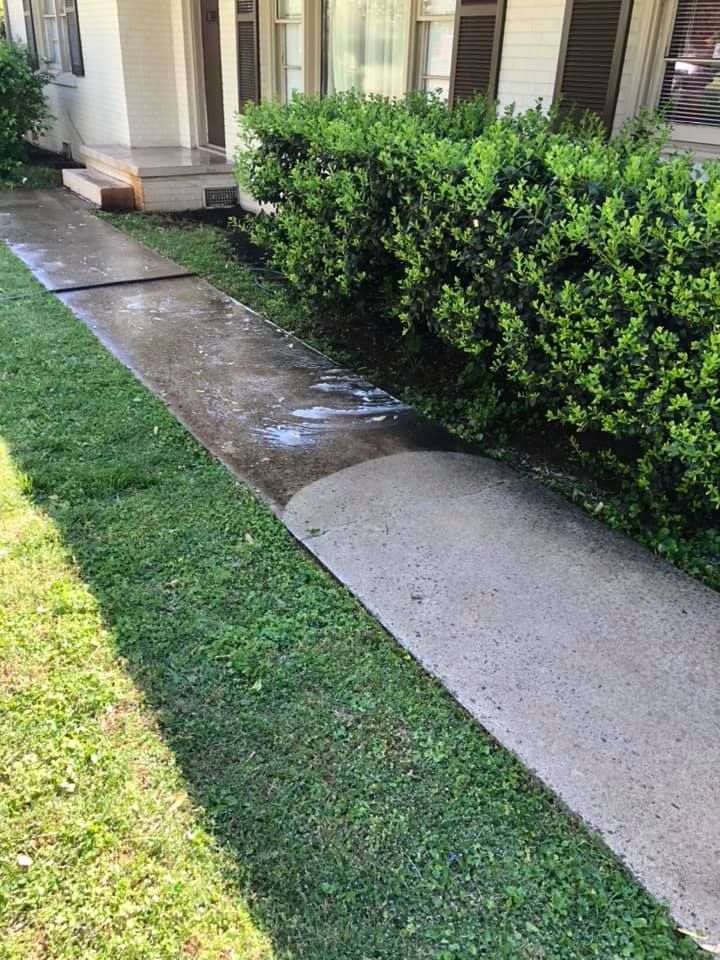 Concrete sidewalk beside a house, bordered by green grass and a trimmed hedge.